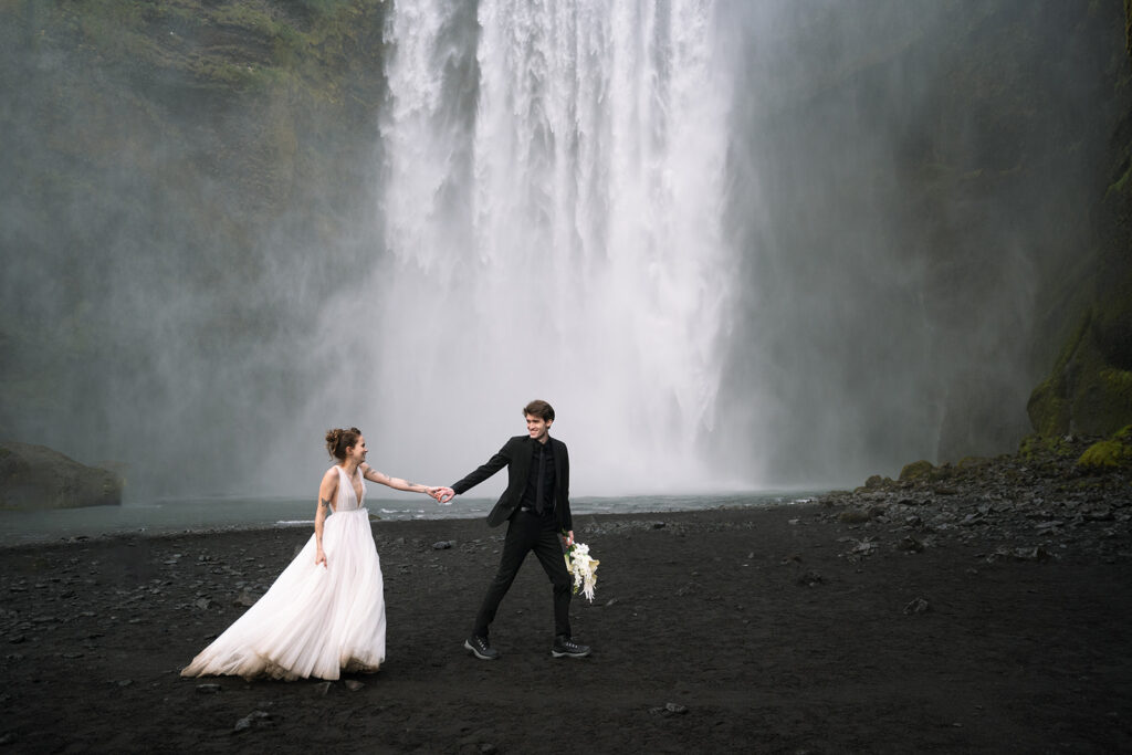 Layla and Kyle holding hands at Skogafoss waterfall during their Icelandic wedding.